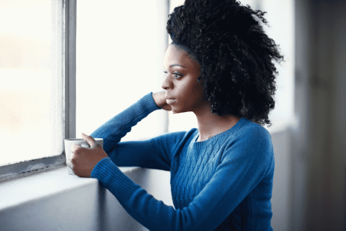 Femme pensive en chandail bleu tenant une tasse de café, regardant par la fenêtre dans un moment de pause au travail.